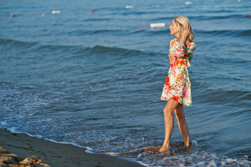 Fototapeta na wymiar Blonde woman wearing dress standing on sand beach at sea shore enjoying view of sunset.