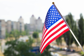 Flag of the United States of America set against blue sky, green trees, city street and multi-storey buildings. US flag display on a house window. The holiday flag of the USA is illuminated by the sun