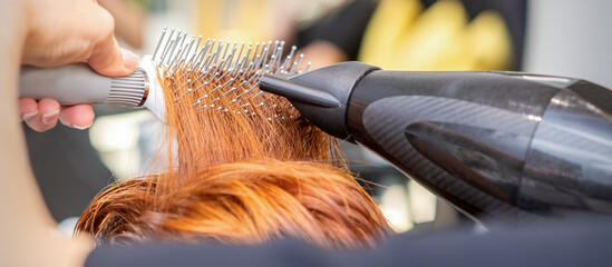 Closeup of master's hand with blow-drying and hairbrush blowing female red hair in a salon