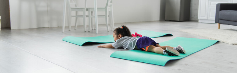 Toddler asian girl in sportswear lying on fitness mat at home, banner