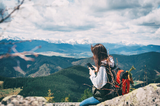 Young woman relaxing on the top mountains. The brunette holds a mobile phone in her hands and takes pictures of the landscape. Side view.