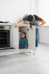 Mother looking at cheerful asian kid in kitchen cabinet