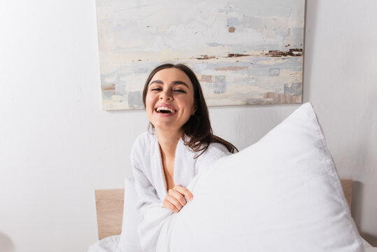 happy young woman in bathrobe holding pillow while looking at camera
