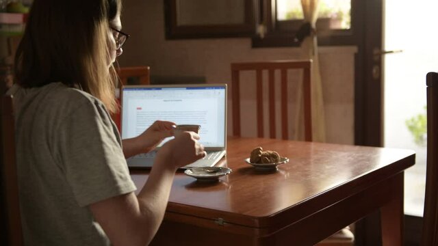4k a girl working with laptop in the kitchen, cessing a work, taking a cup of coffee to break. Domestic life, remote work and study at home, online learning, smartworking, zoom in