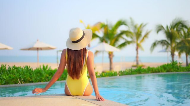 Backside view of the woman sitting on edge of swimming pool at exotic hotel in Hawaii near the beach in yellow monokini and white hat and hitting water with her legs, beach umbrellas and Palms