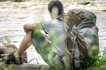 A male traveler with a large hiking backpack sits resting near the river.