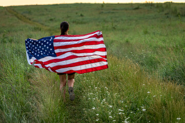 Back view of a girl with an American flag in a field among the grass.