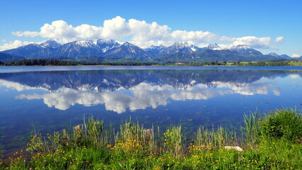malerisches Panorama am Ufer des Hopfensees im Allgäu mit Spiegelung der Wolken im See und Sonne
