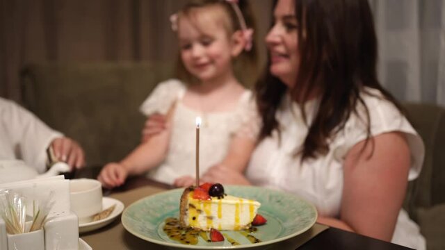 girl with mother and grandmother light and blow candle on piece of festive cake