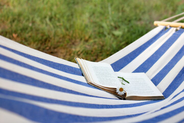 open book with a bookmark of a chamomile flower on a hanging hammock in the garden. outdoor reading in summer