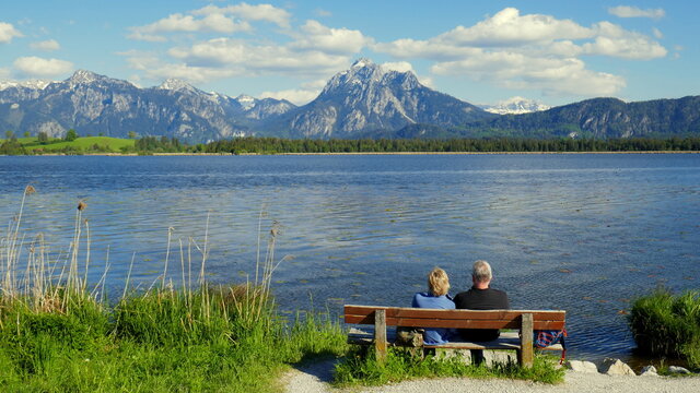 älteres Paar genießt die herrliche Aussicht von einer Bank über den Hopfensee auf die Berge im Allgäu 