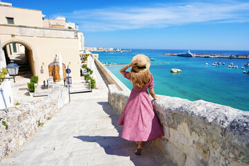 Fototapeta na wymiar Beautiful young woman with hat walking along the ancient walls of Otranto looking at stunning panoramic view of Otranto village in Salento, Italy