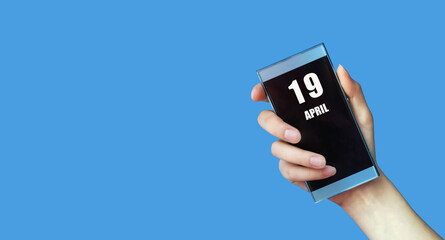 April 19. 19th day of the month, calendar date.Woman's hand holds mobile phone with blank screen on blue isolated background.Spring month, day of the year concept