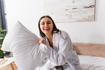 playful young woman in bathrobe holding pillow and laughing while looking at camera