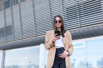 Woman wearing trendy sunglasses walks down the central city street and uses her phone. Pretty summer woman in white jacket walks down the street looking at her mobile phone.