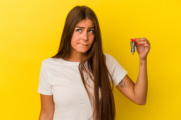 Young caucasian woman holding a home keys isolated on yellow background confused, feels doubtful...