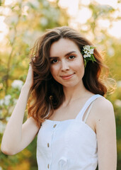 Fototapeta premium A close-up portrait of a happy pretty gentle smiling young woman with a hairstyle in a white cotton dress having fun walking alone enjoying the smell of blooming flowers in a summer green outdoor park