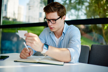 Serious male entrepreneur with business card in hand writing contact information to planner textbook,Caucasian man in optical spectacles for provide eyes protection making notes while learning indoors