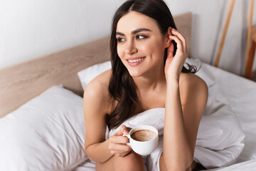 brunette woman smiling while adjusting hair and holding cup of coffee in bed