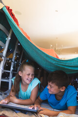 Caucasian brother and sister smiling, lying in blanket fort using tablet at home