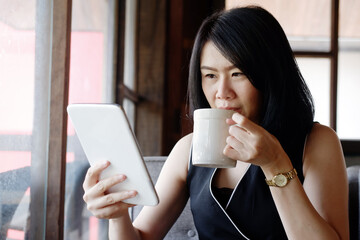 Serious Asian businesswoman is drinking a cup of coffee and holding tablet for online shopping in social media for relaxing at coffee shop. Lifstyle of businesspeople with techonology concept.