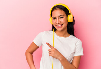 Young Venezuelan woman listening to music isolated on pink background smiling and pointing aside, showing something at blank space.