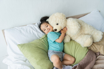Smiling asian kid holding teddy bear on bed