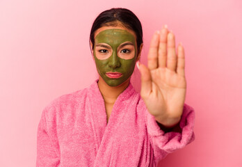 Young Venezuelan woman wearing a bathrobe and facial mask isolated on pink background standing with outstretched hand showing stop sign, preventing you.