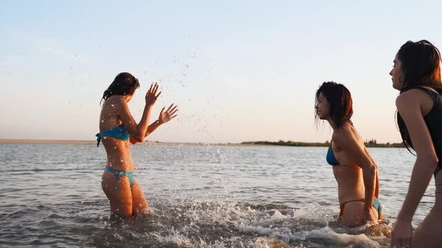 Happy wet girls in bikini run into sea and play splashing water to each other on sunset. Cheerful female friends have fun making splashes in pond. Young women go to swim, bathe in lake. Slow motion.