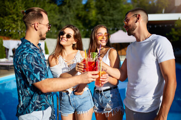 Group of friends having fun at poolside summer party clinking glasses with summer cocktails on sunny day near swimming pool. People toast drinking fresh juice at luxury villa on tropical vacation.