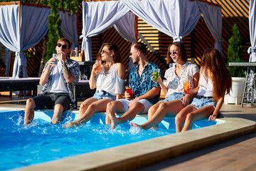 Group of friends having fun at poolside party clinking glasses with fresh cocktails sitting by...