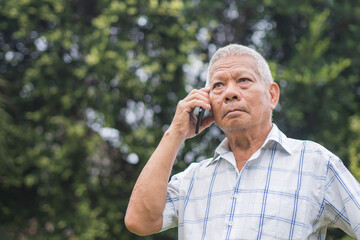 A senior man is using a smartphone looking up and a worried face while standing outdoors in the garden. Space for text. Aged people and communication concept