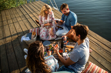 Group of friends having fun on picnic near a lake, sitting on pier eating and drinking wine.
