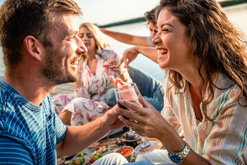 Group of friends having fun on picnic near a lake, sitting on pier eating and drinking wine.
