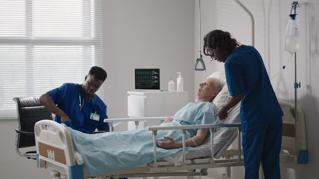 A team of african ethnic group doctors a man and a woman talk to an elderly male patient. A doctor and a nurse talk to a bedridden patient in a hospital ward