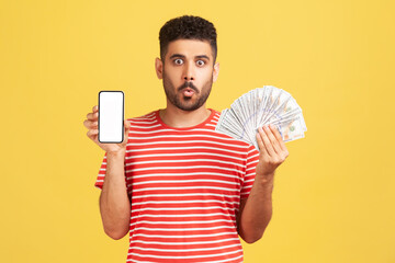 Funny man with beard in red striped t-shirt holding in hands empty display smartphone and credit card, looking at camera with surprised expression. Indoor studio shot isolated on yellow background