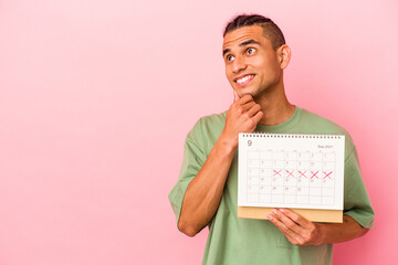 Young venezuelan man holding a calendar isolated on pink background looking sideways with doubtful...