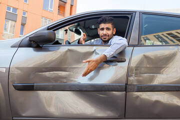 Portrait of confused handsome bearded man wearing blue shirt sitting behind the wheel of dented car with puzzled facial expression, showing dents and scratches of automobile door. Outdoor shot. - obrazy, fototapety, plakaty