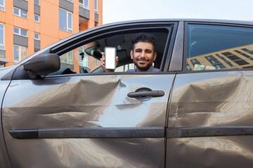 Positive smiling bearded man sitting in dented car and showing mobile phone with empty screen to camera, offers insurance inspection services after road accident. Outdoor shot. - obrazy, fototapety, plakaty