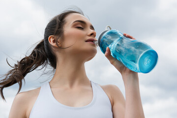 low angle view of young woman in crop top drinking water against blue sky with clouds