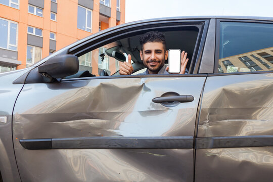Portrait of bearded man in crashed car looking at camera and smiling, showing thumb up and smart phone with blank screen, advertising insurance inspection services after road accident. Outdoor shot. - Powered by Adobe