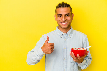 Young venezuelan man holding a cereals bowl isolated on yellow background smiling and raising thumb...