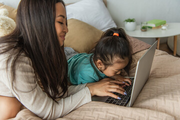Smiling asian woman using laptop near child in bedroom