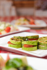 Apetizer from tomatoes, zucchini and mozzarella cheese served on a white plate with other dished on the table.