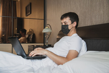 A man is sitting in bed in his bedroom and working at a laptop with a medical mask on his face
