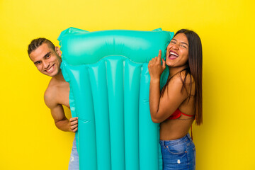 Young latin couple holding an air mattress isolated on yellow background