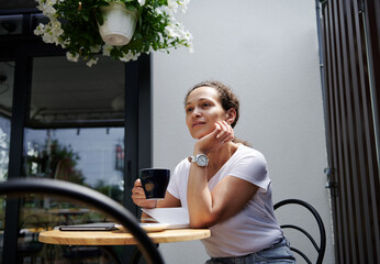 Charming woman sits in a cafe on a summer terrace and looks thoughtfully into the distance with a cup of coffee in her hand