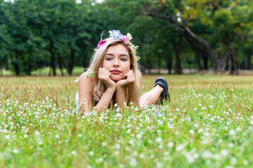 Enjoyment young woman with flower on her hair blowing on dandelion flowers while lying on green grass in the park. Lovely girl blond long hair holding grass flower in her hands at outdoor.