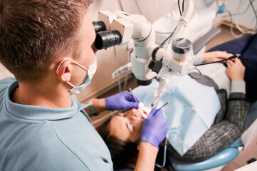 Close up of male dentist in medical mask using dental diagnostic microscope while checking patient teeth. Young woman lying in dental chair at dental office. Concept of dentistry and dental care.