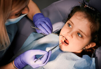 Close up of adorable little girl with open mouth while dentist checking kid teeth, examining child...